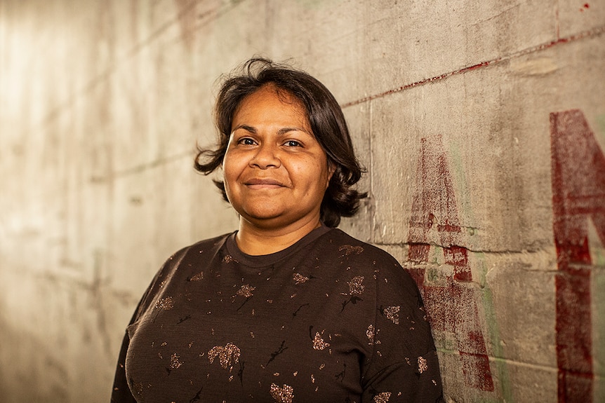 Colour photo of actor and playwright Ursula Yovich smiling and standing in front of faded painted wall.