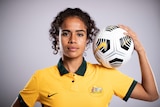 A female soccer player wearing yellow and green holds a ball on her shoulder