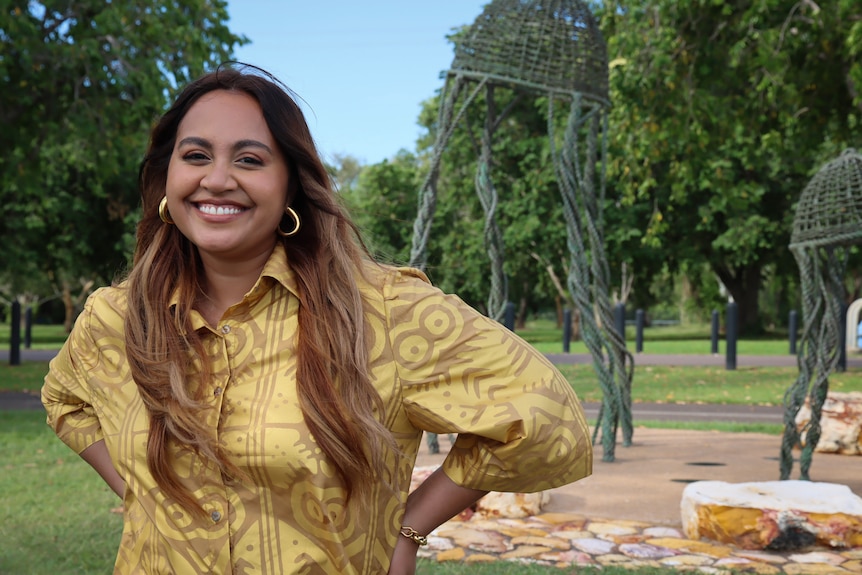 A woman wearing a mustard printed shirt is smiling in a park. Her hair is down and she has her hands on her hips.