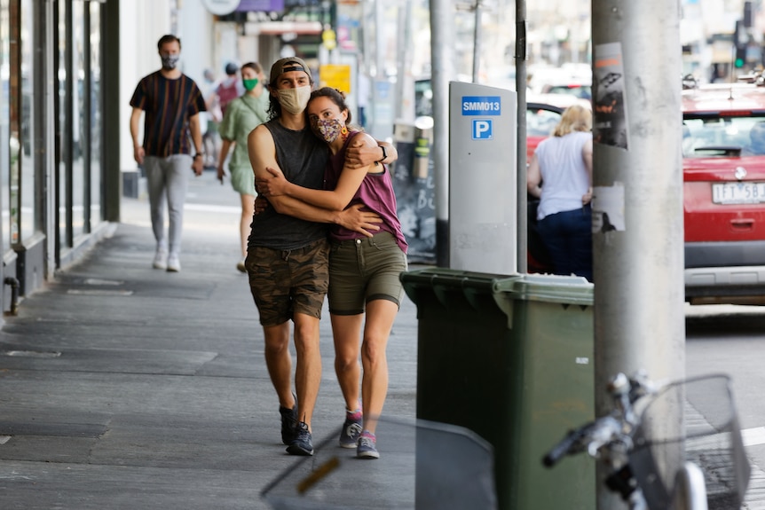 A man and woman, each wearing a mask, hold one another while walking in a city street.