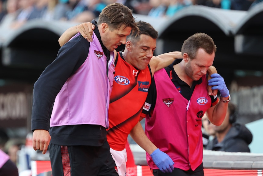 An Essendon AFL player is helped from the field by two members of the club's support staff.