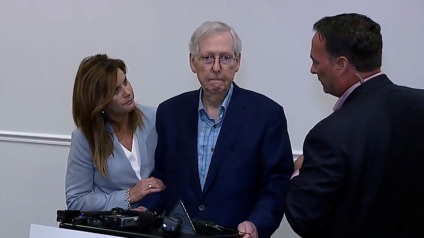 An old man wearing a suit looking stunned and sad being talked to by a man while a woman grabs his arm