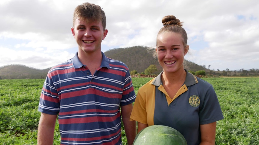A young man and woman stand in a melon field smiling. The woman cradling a melon in her arms.
