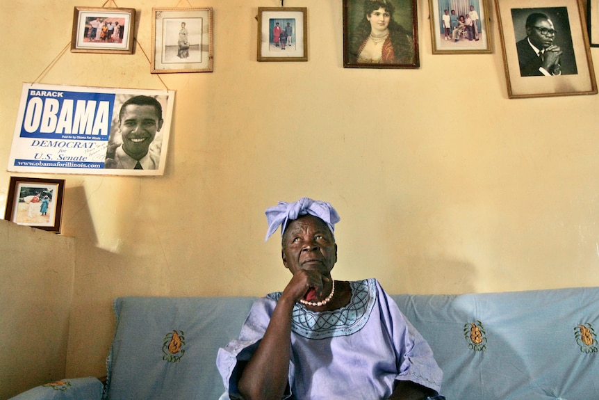 An elderly African woman in purple sits thoughtfuly in a room with an "Obama for US Senate" sign behind her on wall.