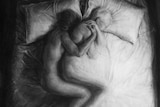 A drawing of a person curled up naked behind their dying partner in a hospital bed.