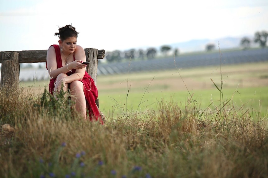 A young lady sits down in a paddock. She wears a red dress, hair and makeup. She looks at her phone.