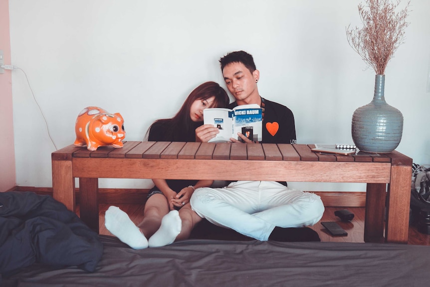 An Asian couple sit side by side at a low table by their bed, reading a book, and considering a shared bedtime.
