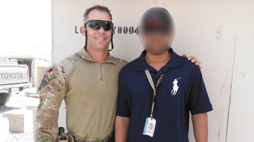 A soldier, in uniform and sunglasses, puts his arm around a man wearing a navy polo shirt. The second man's face is blurred.