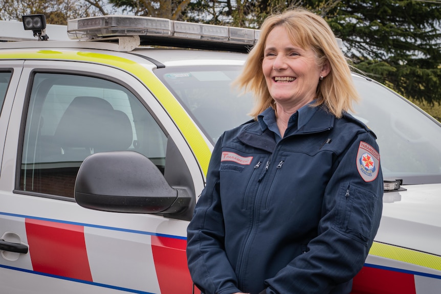 A woman in ambulance uniform stands next to an ambulance vehicle smiling.