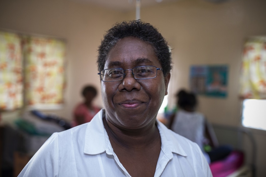 A woman with rectangular glasses smiling in a room with two mother's in the background 