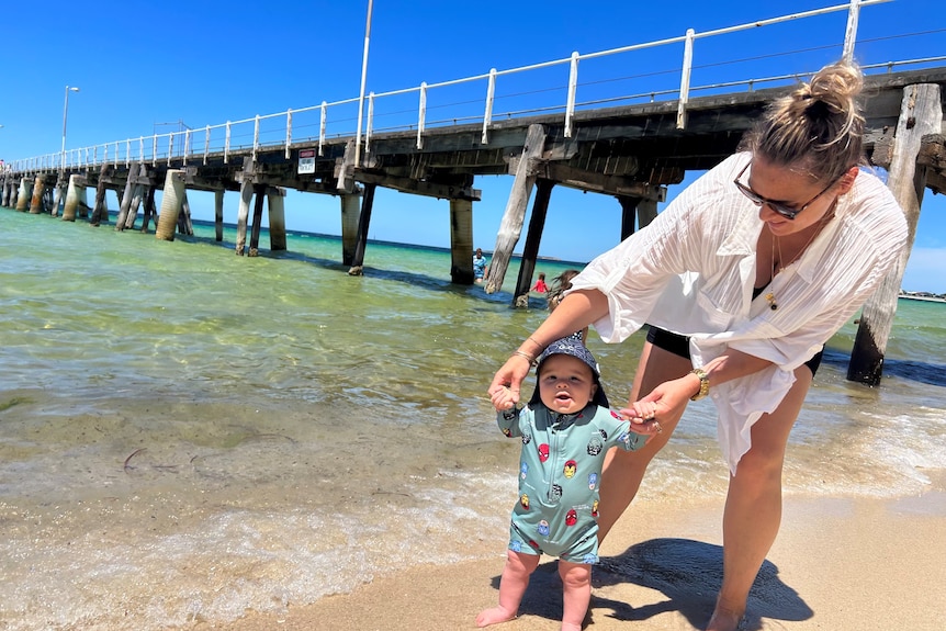 Mum helps little toddler stand at shore of beach