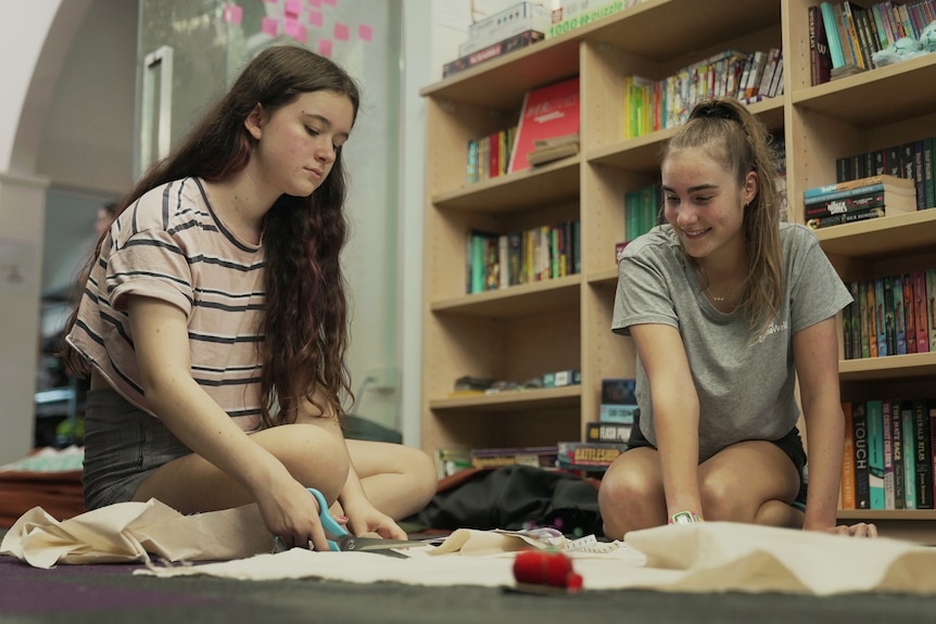 Two teenage girls kneel on the floor, one watching the other cut material.