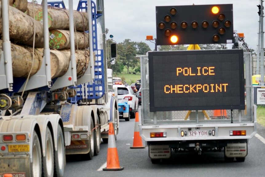 A logging truck waits behind a line of cars alongside a police checkpoint sign.