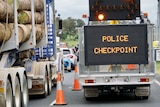A logging truck waits behind a line of cars alongside a 'police checkpoint' sign.