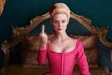 Fanning sits on a bed in a hot pink corseted dress, raising her middle finger
