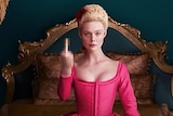 Fanning sits on a bed in a hot pink corseted dress, raising her middle finger