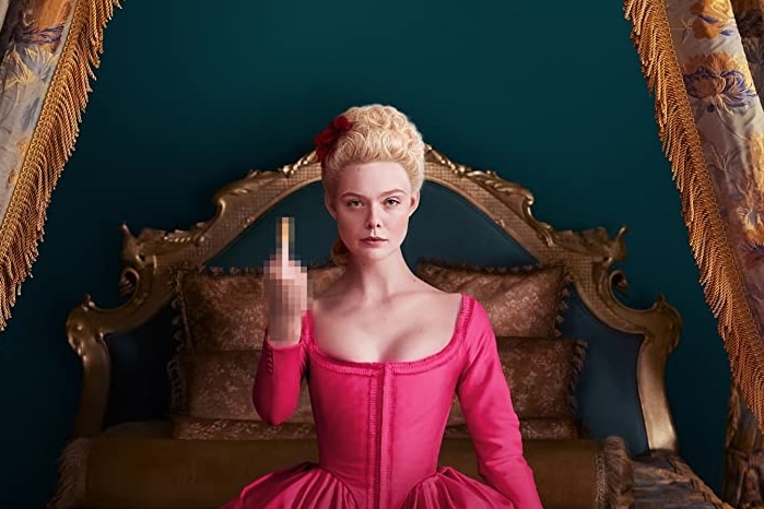 Fanning sits on a bed in a hot pink corseted dress, raising her middle finger