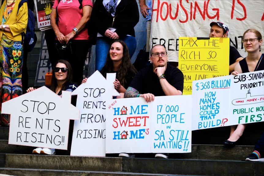 People sitting holding placards with signs about rent rises and housing affordability