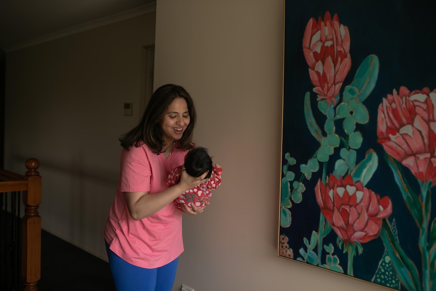 Smiling woman with black hair around shoulders holds baby clad in printed pink onesie, large painting on wall of flower.