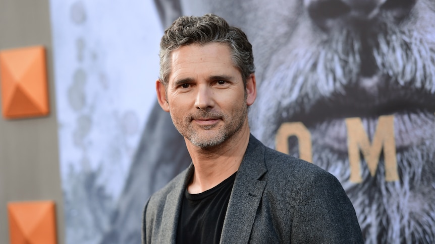 Late-40s man with salt and pepper hair and facial hair, in black tee and grey suit jacket, standing looking at camera.