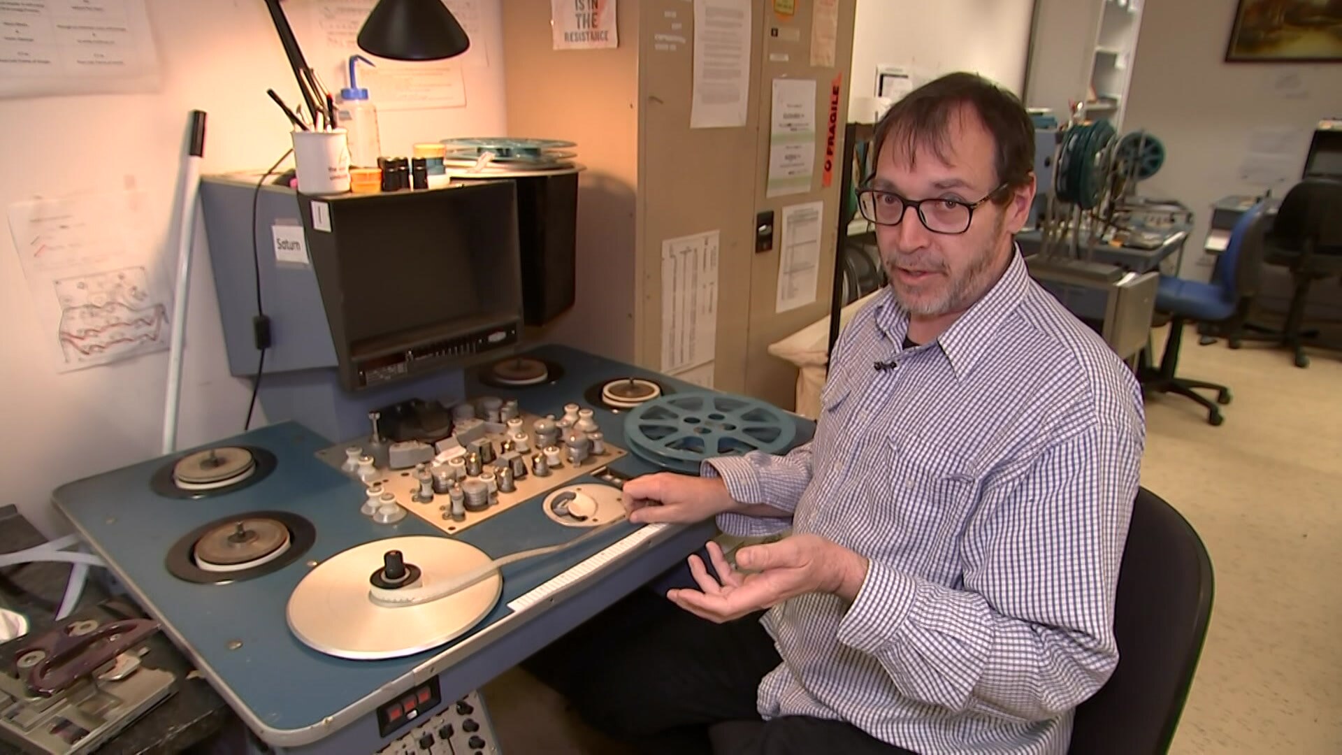 Man sitting at old film machine with film laced up on reels. He is gesturing towards film in mid-conversation.