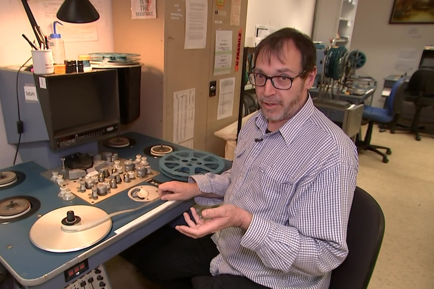 Man sitting at old film machine with film laced up on reels. He is gesturing towards film in mid-conversation.