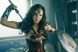 Screen from film shows Wonder Woman (Gal Gadot) in an action pose pulling a sword from behind her and holding a shield.