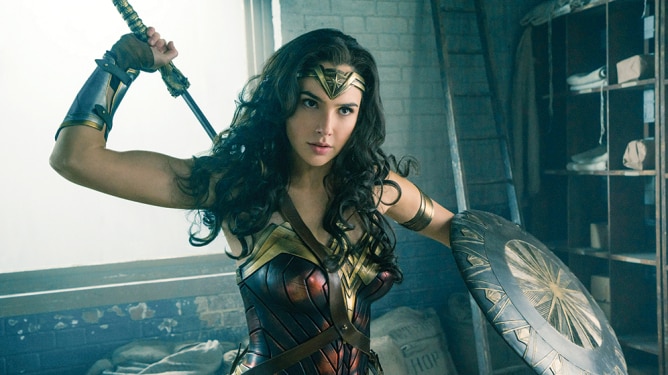 Screen from film shows Wonder Woman (Gal Gadot) in an action pose pulling a sword from behind her and holding a shield.