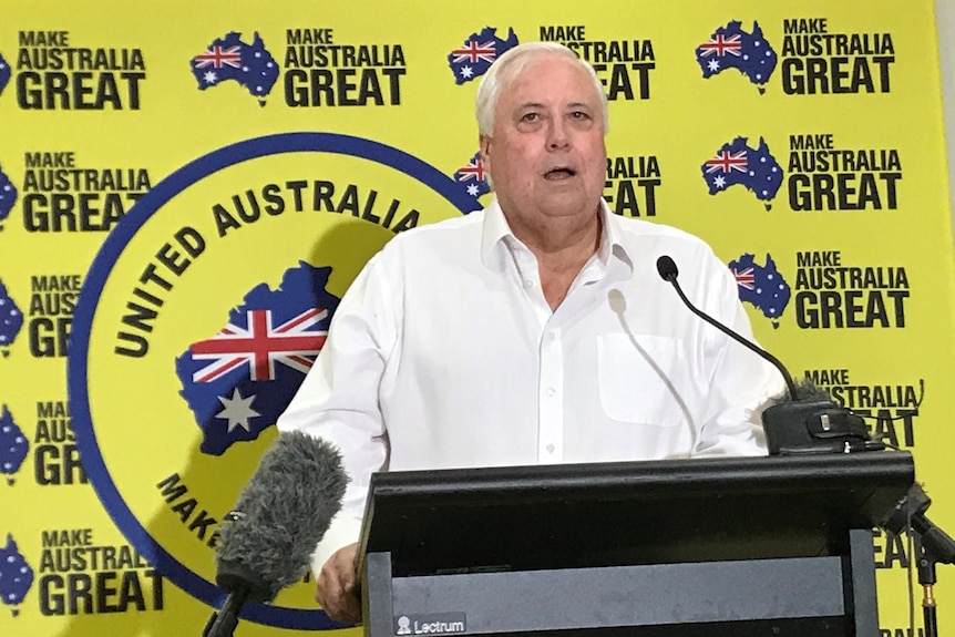 An elderly man in a white shirt speaks at a podium with yellow political sign behind.