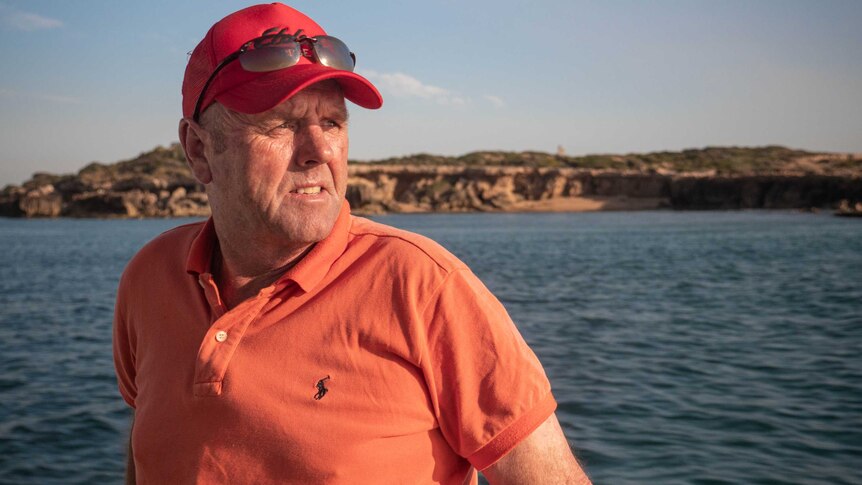 John Fenton on the side of a boat, wearing orange t-shirt and red cap, water and sea cliffs behind him.