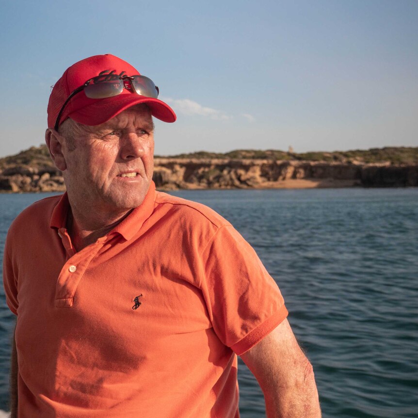 John Fenton on the side of a boat, wearing orange t-shirt and red cap, water and sea cliffs behind him.