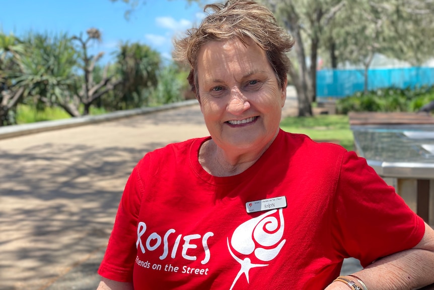 Woman with short hair sitting at bench smiling, wearing red t-shirt that reads "Rosies" with depiction of white rose
