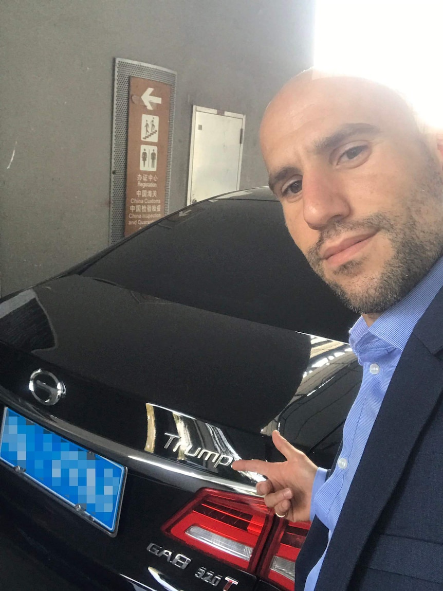 Stavro D'Amore, with bald head and facial hair, takes a selfie in front of a black sedan.