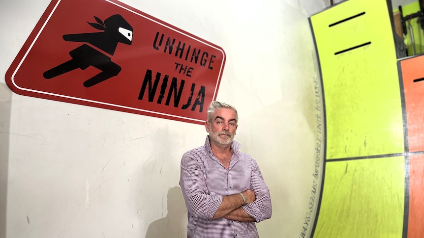 A man stands with crossed arms in front of a steep ramp and a sign reading 'Unhinge the Ninja'.