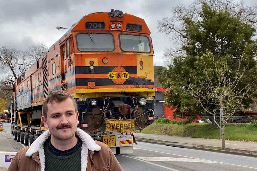 A man stands in front of a orange diesel train engine travelling on the back of a semi trailer