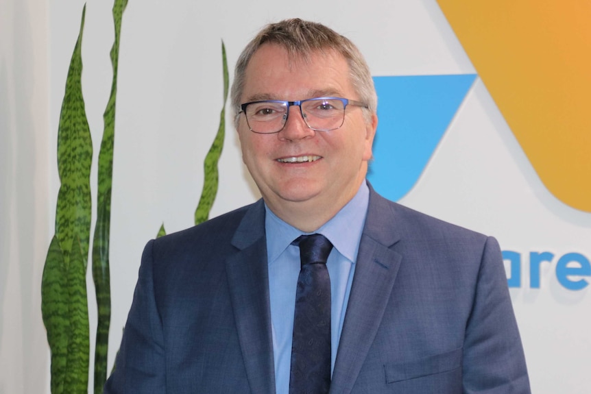 A head and shoulders shot of a smiling Anglicare CEO Mark Glasson posing for a photo indoors wearing a blue suit and tie.