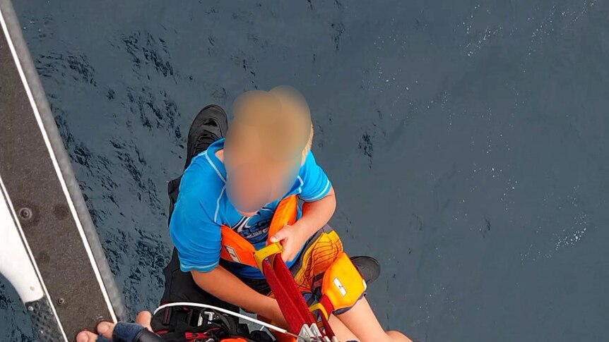 A helicopter rescue officer winches a small boy on to the aircraft, with ocean below.