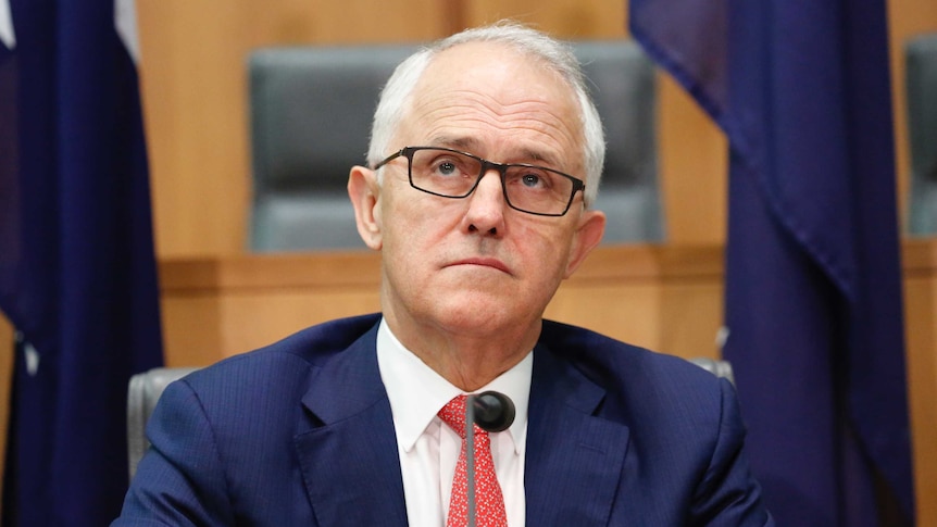Turnbull wearing a blue suit, red tie and dark-rimmed glasses, looks skyward with a sombre expression.