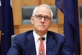 Turnbull wearing a blue suit, red tie and dark-rimmed glasses, looks skyward with a sombre expression.