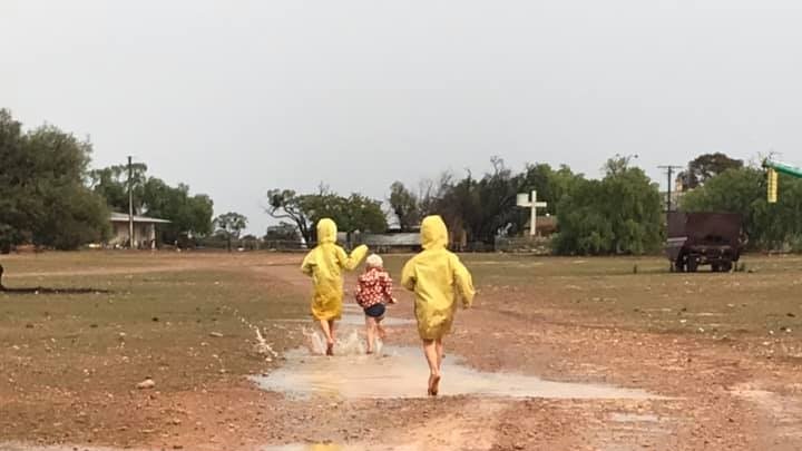 Three kids in raincoats play in puddles in the mud after recent rainfall.