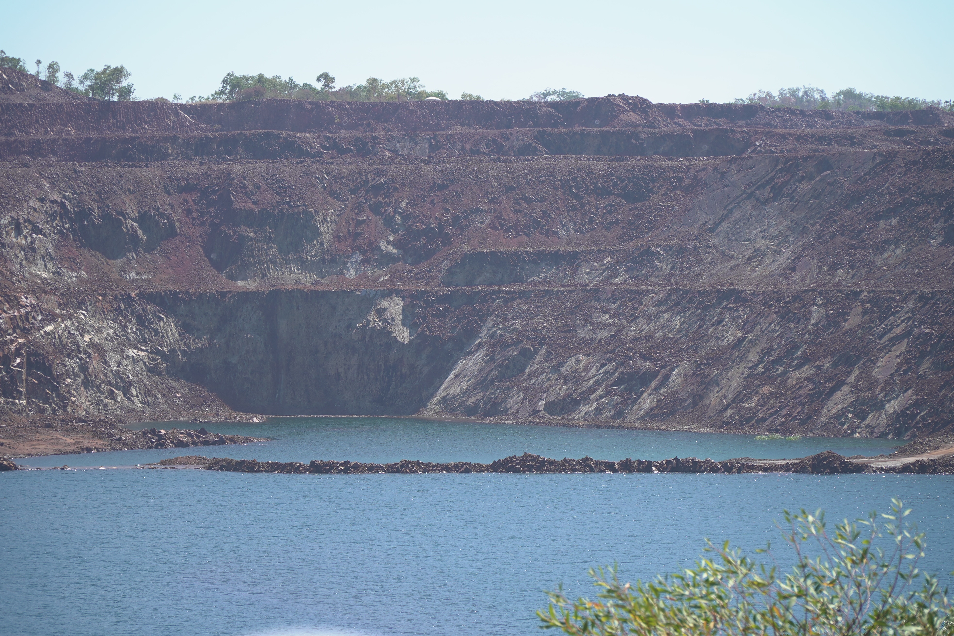 A section of a wall of an open-cut mining pit mostly full of blue water.