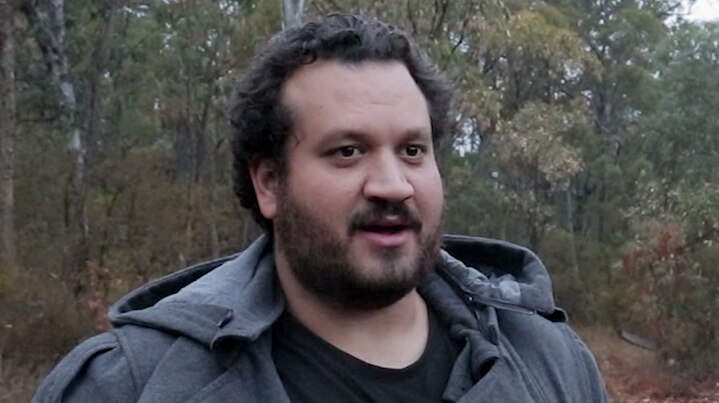 An Aboriginal man stands on a rock in the bush on a cloudy day, facing a reporter.