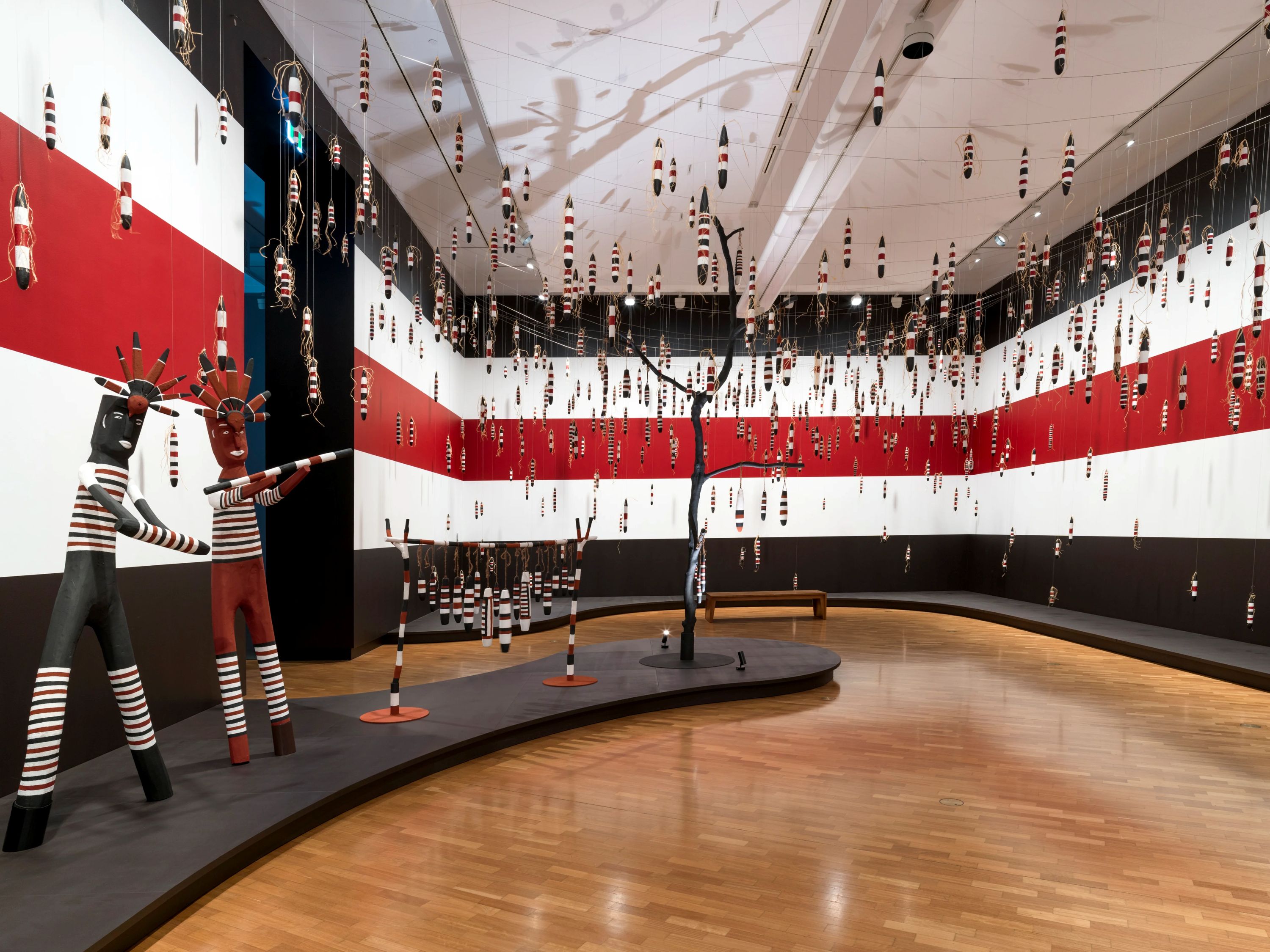 En una galería, tallas cilíndricas de madera pintada cuelgan del techo, junto a figuras escultóricas de madera y un árbol en reposo.