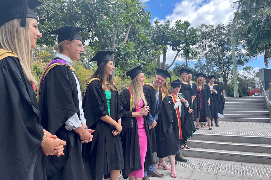  A group of university students standing in a line along some steps, wearing black graduation robes.