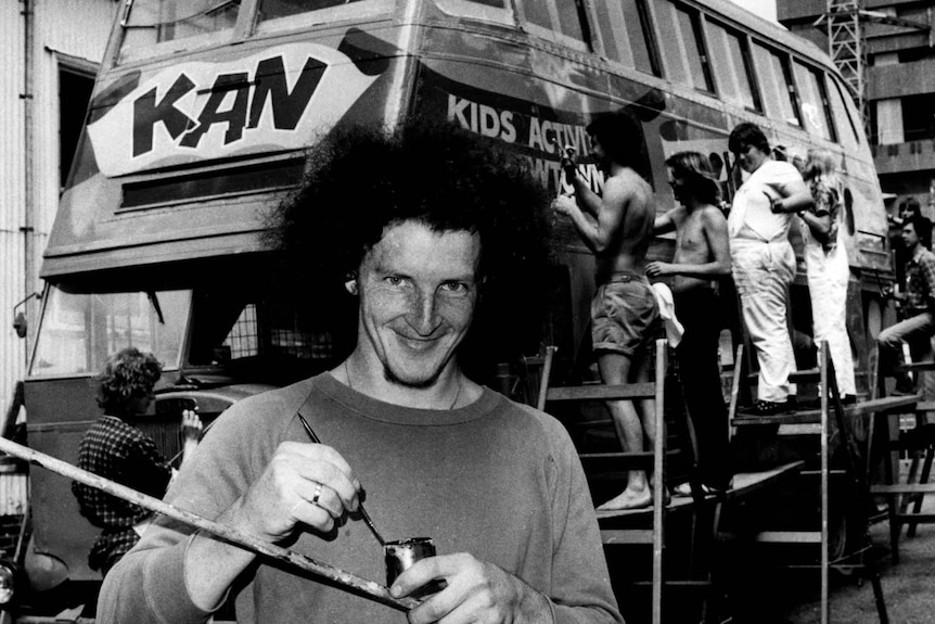 A black and white image of a young man with frizzy hair smiling in front of a bus being painted, holding a paintbrush.