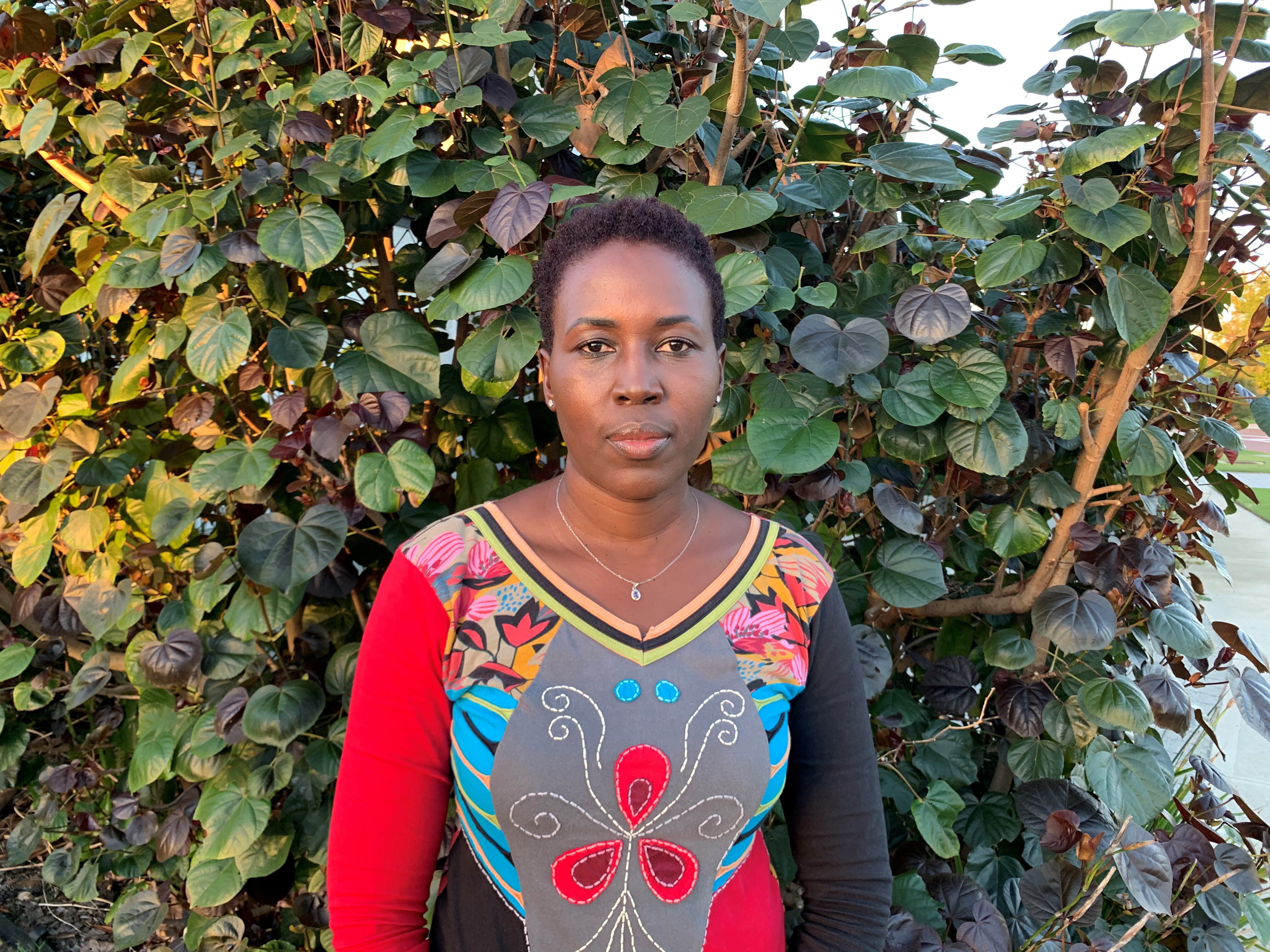 Barbara wearing a colorful top, standing in front of green bushes in a front garden.