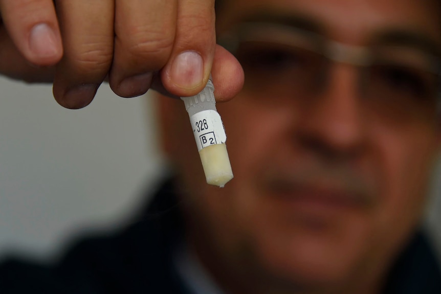 A man out of focus hold a vial with a cream colour substance in it