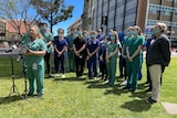 A group of hospital stuff wearing scrubs stand on a lawn, with one in front of microphones.