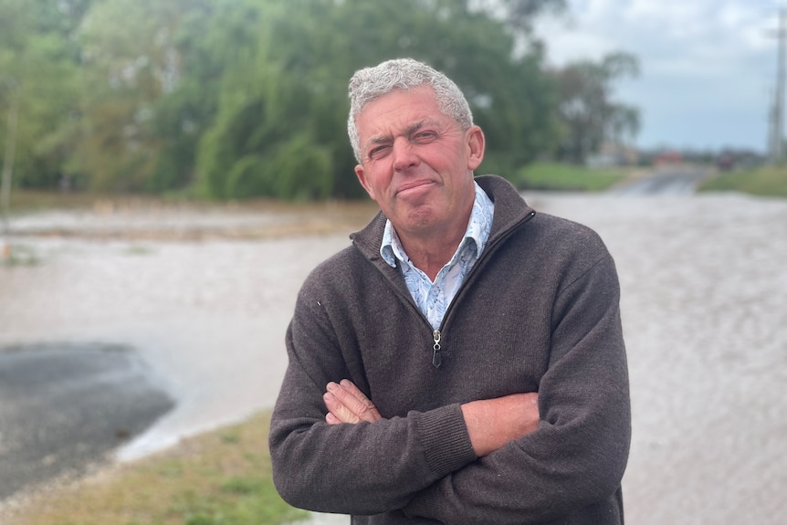 An older man with grey hair stands in front of a waterway with his arms folded.