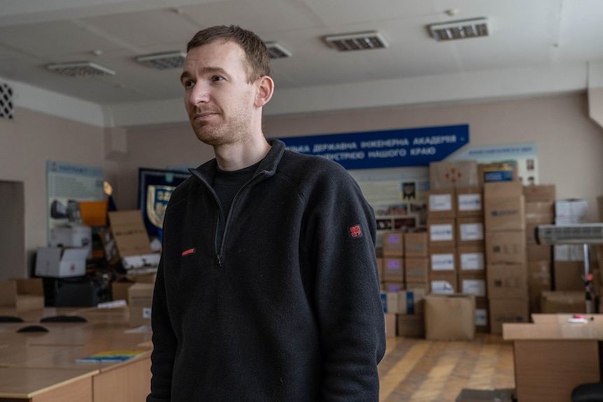 A skinny man in a jumper looks to the left as he stands in a room full of aid supplies